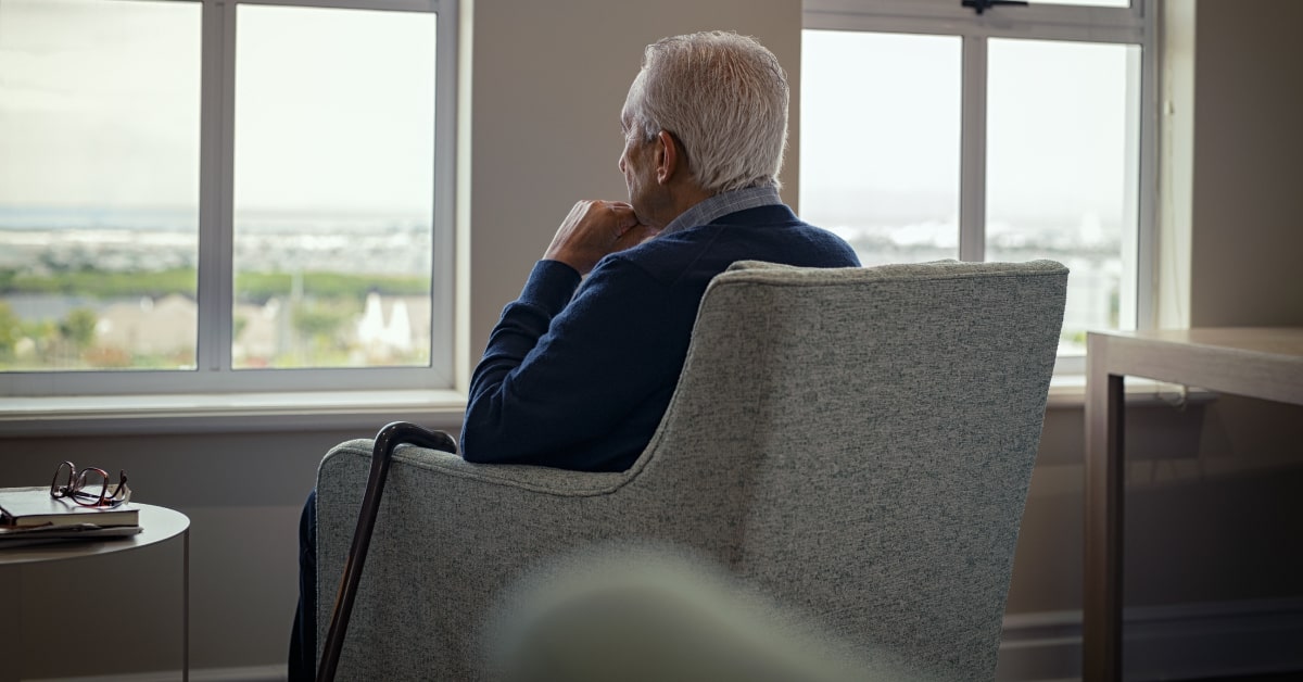 Hombre mayor sentado de espaldas, mirando por la ventana pensativo
